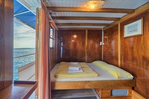 Kompakte Holzkabine auf Schiff Linda mit Doppelbett, gelber Bettwäsche, weißen Handtüchern, Bullauge mit Meerblick und Schiffsbild.