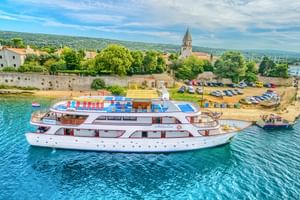 Weißes Kreuzfahrtschiff Andela Lora vor Anker in türkisfarbenem Wasser nahe kroatischer Küstenstadt mit Steingebäuden und Kirchturm.