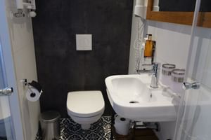 Compact bathroom on MS MAGNIFIQUE IV with white wall-mounted toilet, sink with chrome faucet, patterned floor tiles, and dark accent wall.