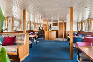 Geräumiges Restaurant an Bord der Floor van Aemstel mit Holztischen, bunten Sitzgelegenheiten, gemusterten Bänken und Fenstern an beiden Seiten.