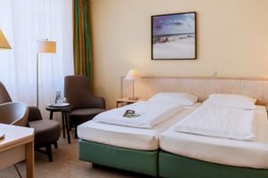 Helles Hotelzimmer mit zwei Einzelbetten, grünem Sockel, Holzkopfteil, Sitzbereich mit braunen Sesseln, Stehlampe und Landschaftsbild an beiger Wand.