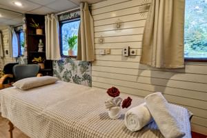 Massageliege mit weißer Wäsche und roten Rosen an Bord der MS Prinzessin Katharina. Raum mit weißer Holzverkleidung, Fenstern und Pflanzen.