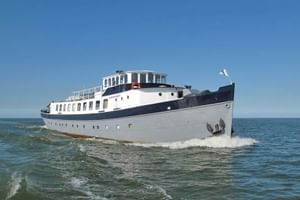 Weiß-dunkelblaues Passagierschiff Floor van Aemstel fährt auf ruhigem Wasser unter klarem blauen Himmel, mit weißer Flagge am Heck.