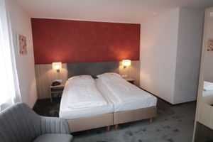 Modernes Hotelzimmer mit zwei Einzelbetten, roter Akzentwand, Holzkopfteil, Nachttischlampen, grauem Sessel und Fenster mit weißen Vorhängen.