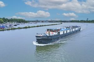 MS Magnifique IV, a white and blue river cruise ship, sailing on a wide river with marina and green landscape in the background.