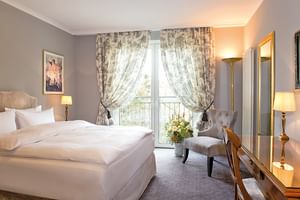 Elegantes Superior-Doppelzimmer mit beigem Bett, grauem Sessel, gemusterten Vorhängen, Balkontüren und warmer Beleuchtung im Victors Residenz Hotel.