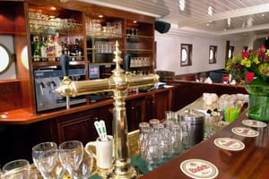 Elegante Schiffsbar mit Messingbierzapfhahn, Gläsern, Flaschen in Regalen, weißer Decke, Bullaugen und frischen Blumen auf polierter Holztheke.