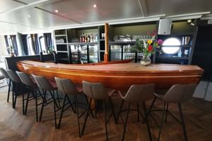 Modern restaurant bar on MS Magnifique IV with curved wooden counter, gray bar stools, and glass-front refrigerators displaying beverages.