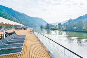 Sonnendeck der MS Prinzessin Katharina mit Holzboden, Sonnenliegen und weißem Geländer. Flusslandschaft mit bewaldeten Bergen im Hintergrund.