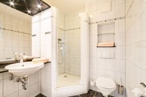 Modernes Hotel Seeblick Badezimmer mit weißen Fliesen, wandmontiertem Waschbecken, Glasduschkabine, WC und dunklen Bodenfliesen.