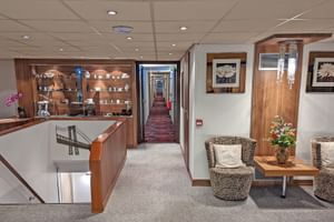 Rezeption der MS Normandie mit Holztheke, Regalen, Korbstühlen mit Kissen und einem Korridor zu den Kabinen.