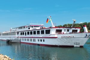 Weißes Flusskreuzfahrtschiff MS Normandie mit rotem Zierstreifen und deutscher Flagge auf ruhigem Wasser, grünes Ufer im Hintergrund.