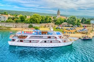 White cruise ship MY Andela Lora moored in turquoise waters near a Croatian coastal town with stone harbor walls, church tower, and parked cars.