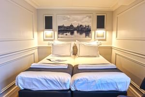 Elegante Doppelkabine auf der MS Magnifique Vier mit zwei Einzelbetten, weißer Bettwäsche, Wandleuchten und gerahmtem Stadtbild über dem Kopfteil.