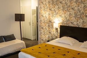 Hotelzimmer im Grand Hotel Toulouse mit zwei Betten, schwarzen Kopfteilen, gelber Decke, Blumentapete, Stehlampe und Eingang zum Bad.