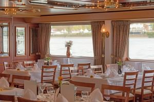 Eleganter Speisesaal auf der MS Switzerland mit gedeckten Tischen, Holzstühlen, weißen Tischdecken und großen Fenstern mit Flussblick.