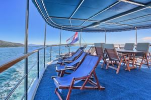 Sonnendeck des Schiffs San Snova mit blauen Liegestühlen und Holzstühlen unter Sonnensegel. Kroatische Flagge, türkisfarbenes Meer und Küste.