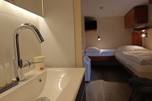 Modernes Badezimmer auf MS Fluvius mit Chromarmatur und weißem Waschbecken, Blick in Kabine mit zwei Betten und Holzmöbeln.