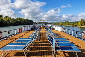 Sonnendeck der MS Olympia mit Reihen blauer Liegestühle auf Holzdeck. Flusslandschaft mit grünen Bäumen und blauem Himmel mit Wolken im Hintergrund.
