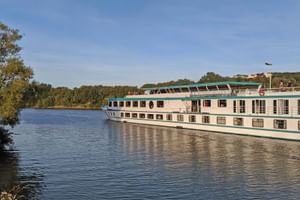 Flusskreuzfahrtschiff MS Switzerland mit weißem Rumpf und grünen Akzenten auf ruhigem Fluss, umgeben von Bäumen unter blauem Himmel.