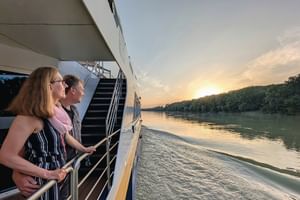 Zwei Passagiere auf dem Deck des Flusskreuzfahrtschiffs SE Manon beobachten den Sonnenuntergang über der Donau mit bewaldeten Ufern.