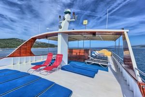 Sonnendeck des Kreuzfahrtschiffs Andela Lora mit blauen und roten Liegestühlen unter gebogenem Holzdach, mit Blick auf blaues Meer.