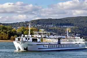White river cruise ship MS Veloria sailing on the Rhine with forested hills and a town visible in the background under a partly cloudy sky.