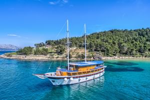 Weiße Segelyacht San Snova mit zwei Masten vor Anker in kristallklarem türkisfarbenem Wasser nahe einer bewaldeten Küste unter blauem Himmel.
