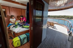 Innenansicht der Andela Lora Schiffskabine mit zwei Gästen auf einem Bett mit Gepäck, Holzvertäfelung und offener Tür zum sonnigen Deck.