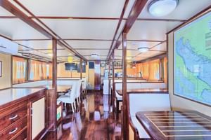 Innenraum des San Snova Schiffssalons mit polierten Holzböden, weißen Esstischen, Seekarten an den Wänden und orangefarbenen Vorhängen.