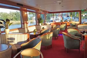 Innenraum der MS Fluvius Lounge mit grünen Sesseln, gestreiften Sofas und runden Tischen auf rotem Teppich. Große Fenster zeigen Fluss und Boote.