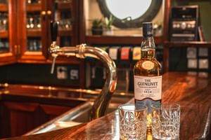 The Glenlivet 1824 Whiskyflasche auf Holztresen mit zwei Kristallgläsern und Bierhahn, Vitrinen im Hintergrund sichtbar.