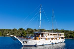 Weiße Segelyacht Magellan mit zwei Masten vor Anker in ruhigen blauen Gewässern nahe einer bewaldeten Küste unter klarem Himmel.