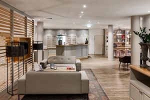 Moderne Hotellobby mit grauen Sofas, Holzraumteiler, Rezeptionstresen und Zierpflanzen. Heller Holzboden im gesamten Raum.