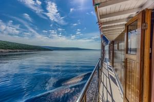 Blick von Deck 3 des Schiffes Linda mit Holzgeländer, Kabinentüren und blauem Meerwasser mit bewaldeter Küste unter bewölktem Himmel.