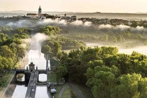 Luftaufnahme von Mělník mit Kirchturm über Morgennebel. Schleuse an der Elbe im Vordergrund, umgeben von grünen Wäldern.