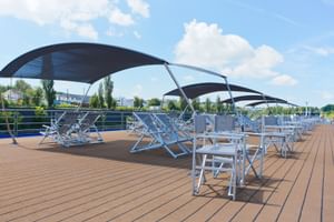Sonnendeck mit Holzboden mit blau-weißen Klappliegen unter schwarzen Sonnendächern. Metallgeländer säumen das Deck, Bäume im Hintergrund sichtbar.