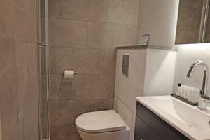 Modernes Badezimmer auf der MS Fluvius mit beigen Fliesen, Glasdusche, weißem WC und dunklem Waschtisch mit weißem Waschbecken.
