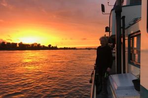 Person standing on deck of MS Flora cruise ship during golden sunset on Mosel River, with orange sky reflecting on water and shoreline visible.