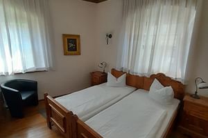 Doppelzimmer in der Schlossschänke Bautzen mit geschnitzten Holzbetten, weißer Bettwäsche, Holzboden, Gardinen und schwarzem Sessel.