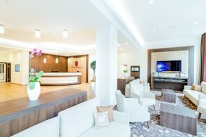 Moderne Hotel Seeblick Rezeption mit geschwungenem weißen Tresen, Holzverkleidung, weißer Sitzbereich mit TV und Orchidee in weißer Vase.