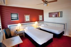 Modernes Hotel-Zweibettzimmer mit zwei Einzelbetten, roter Akzentwand, weißen Möbeln, Schreibtisch mit Stuhl und Deckenventilator.