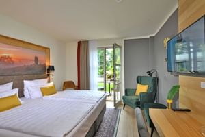 Helles Hotelzimmer mit Doppelbett, gelben Kissen, grünem Sessel, Wandfernseher und Balkontür im Landhotel Schnuck in der Lüneburger Heide.