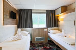 Helle Zweibettkabine auf dem Oberdeck der MS Normandie mit zwei Einzelbetten, holzgetäfelten Wänden, Fenster mit grauen Vorhängen, Wandfernseher und gemustertem Teppich.