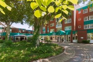 Außenansicht des Upstalsboom Hotels Emden mit roter Backsteinfassade und grünen Akzenten. Gepflasterter Innenhof mit alten Bäumen und Terrasse.