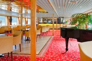 Geräumiger Salon an Bord der MS Normandie mit rotem gemustertem Teppich, Flügel, beigen Sesseln, Holzsäulen und Fenstern mit orangen Vorhängen.