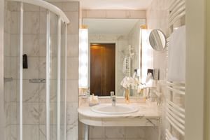 Luxuriöses Marmorbad mit gebogener Glasdusche, weißem Waschtisch, rundem Spiegel und warmer Beleuchtung im Victors Residenz Hotel.