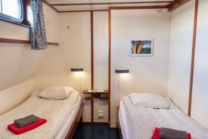Compact twin cabin on MS Sarah with two single beds, reading lights, porthole with curtains, and red towels on each bed.