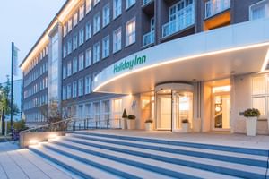 Modernes Holiday Inn Hotelgebäude in Dresden mit beleuchteter Eingangshalle, geschwungenem Vordach, gläserner Drehtür und breiten Steinstufen zum Eingang.