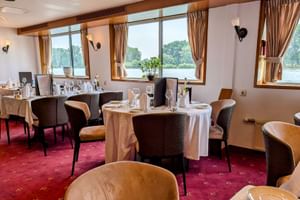 Elegantes Restaurant an Bord der MS Olympia mit weiß gedeckten Tischen, braunen Stühlen und großen Fenstern mit Blick auf einen Fluss.